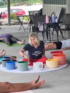 People laying on mats and chairs, woman sits in middle of them using singing bowls
